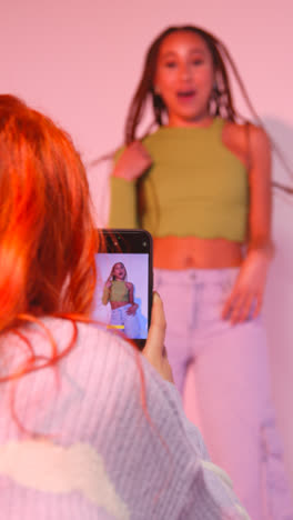 Vertical-Video-Studio-Shot-Of-Woman-Taking-Photo-Of-Friend-Dancing-On-Mobile-Phone-Against-Pink-Background